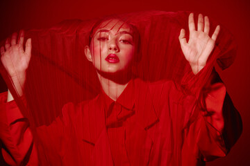 Fashionable young woman in red outfit behind red mesh fabric. Creative beauty concept with strong color contrast on a red background. Artistic high fashion studio shot.