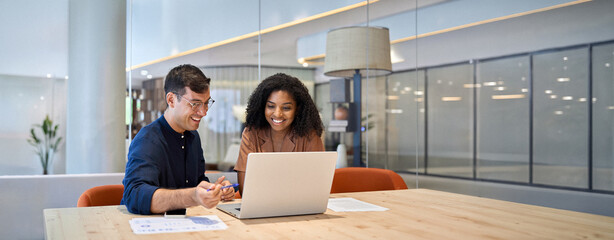 Smiling team professional employees working together on computer discussing online technology data solutions at work. Two young busy diverse coworkers business people using laptop at office meeting.