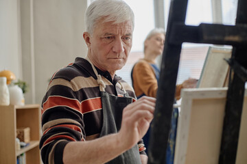 Senior Caucasian man painting on canvas in classroom setting, focused on brushwork, participating in art education activity with older Caucasian woman painting in background