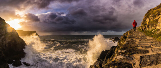 Dramatic Ultrawide Landscape of Powerful Ocean Waves Crashing Against Rocky Cliffs at Sunset with a Person in a Red Jacket Walking Along a Stone Path Under Dark Stormy Clouds on the Coastline.
