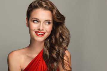 A slender and elegant woman with a beautiful smile. She is wearing a red dress and red lipstick. The girl's hair is styled in a wavy hairstyle. Curly hair, cosmetology, beauty, and makeup.