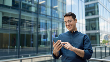 Happy young Hispanic business man manager using financial banking mobile apps on cell phone standing in city, professional businessman holding smartphone in hands looking at cellphone outdoor office.