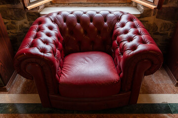 A vintage red leather tufted armchair in Chesterfield style sits beneath an open window against a stone wall, evoking a cozy, classic and nostalgic atmosphere in a warm rustic interior setting