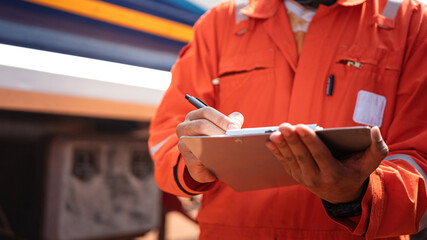 An engineer is checking on chemical hazardous material checklist to verify the safety condition with chemical road tanker as background. Industrial waorking scene, close-up and selective focus.