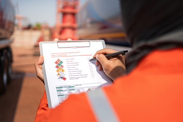 An engineer is checking on chemical hazardous material checklist to verify the safety condition with chemical road tanker as background. Industrial waorking scene, close-up and selective focus.
