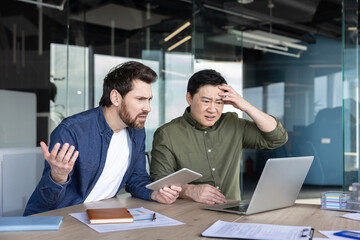 Two businessmen at a modern office desk react with frustration and stress as a laptop error disrupts work, showing confusion, concern, and urgent problem-solving under deadline pressure