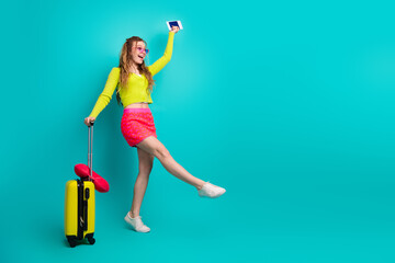 Stylish young woman holding passport and luggage poses in trendy outfit with vibrant teal background for travel theme