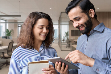 Diverse friendly coworkers colleagues indian male manager and latin female employee talking in office using tablet device, having fun and laughing together discussing project.