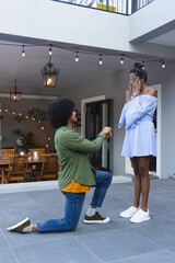African American couple kneeling and proposing on tiled patio under string lights, ring box visible