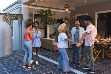 Diverse friends chatting, holding hands on patio with string lights, wicker sofa, wood table