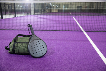 Green duffel, black paddle resting on purple turf by net, white line, glass, lights, copy space