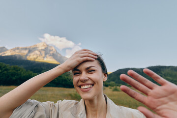 Cheerful woman outdoors, smiling with hand on head, sunlit moment, relaxed posture, natural colors, meadow and mountains in the background, gentle breeze, blue sky