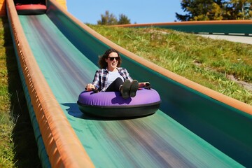 Woman smiles while sliding down tubing track on sunny day. She rides inflatable ring down green slope with grassy sides. Woman wears sunglasses and casual clothes, enjoying fun activity at resort.