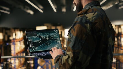 Male army soldier ensuring surveillance in secure military warehouse, personnel monitoring drone dashboard on laptop. Defense logistics to ensure supply chain for military base operations.