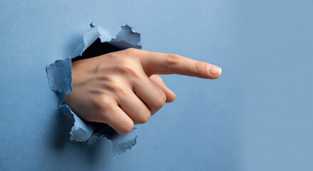 Woman hand pointing finger through a torn blue paper hole. Concept of direction, choice and discovery. Symbolic gesture of communication and information highlighted by a rip in background.