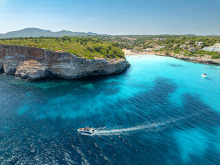 Aerial view of motor boat near resort beach in Mallorca, Spain. Top view of Luxury speedboat cruising in turquoise sea water bay, sandy beach, hotels on high rock cliffs in summer. Drone landscape