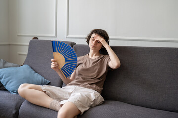 Woman at home suffering from overheating, sitting on sofa in living room holding hand paper fan for cooling down. Tired young female on couch with sick expression, overwhelmed by summer heat or fever