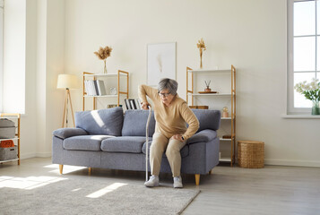 Portrait a of senior gray-haired elderly injured woman getting up from the couch with the help of her crutch in living room at home. Mature female person using walking cane indoors.