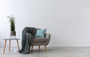 Modern boho cozy corner of home in living room. Gray vintage armchair with blue pillow and blanket, table with green plant in pot, wooden floor, on light wall background, copy space, mockup, nobody