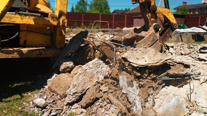 Excavator bucket digging into a pile of broken concrete, rebar, and rubble during demolition work on a construction site under a clear blue sky, showing urban renewal activity