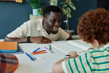 Medium shot of Black male teacher guiding schoolboy during writing practice at desk, supporting literacy and learning. Useful for education, tutoring, child development campaigns