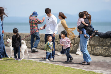 Family enjoys seaside walk with children and dog. Parents smile, guiding kids along stone path. White dog wags tail, walking beside leash. Cloudy sky meets calm ocean horizon. Perfect family moment