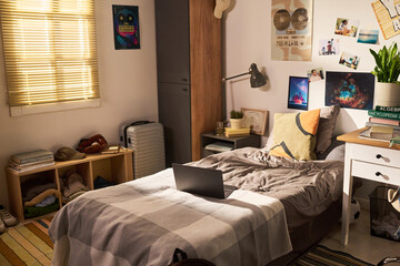 Wide angle shot of student bedroom featuring laptop on bed, study desk, books, suitcase, personal decor, suggesting college living, remote learning, youth lifestyle marketing