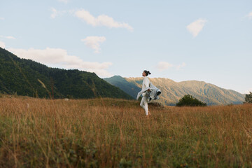 Woman walking in a grassy field near mountains under a blue sky with clouds. Lifestyle concept with nature, freedom, and outdoor activity in soft natural light.