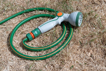 Dried-up, sunburnt grass which has turned yellow from a lack of water, with green hose pipe with grey watering nozzle laying on the ground. The resulting condition of a British garden lawn due to the 