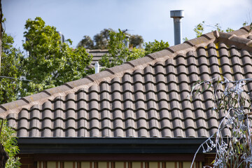 Terracotta tiled roofline of a suburban home with a metal flue vent, guttering, and mature trees in the background. Roof of a residential house in Melbourne, Australia. Concept of roofing, maintenance