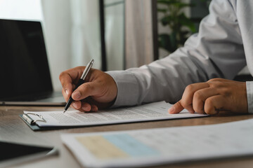 Business person reviewing a printed document with a pen on a clipboard beside a laptop, showing an office document checking process for paperwork verification and administrative review.