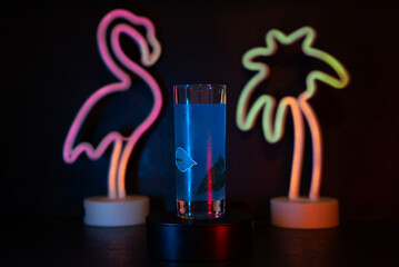 Neon flamingo glows softly beside palm tree. Blue drink sits cool on dark stand. Vibrant lights cast colorful shadows on dark surface. Perfect for tropical, nightlife, or modern bar scenes