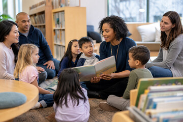 Diverse Children and Parents Listening Together as a Community Volunteer Shares a Picture Book inside a Warm Neighborhood Public Library during a Weekend Story Circle