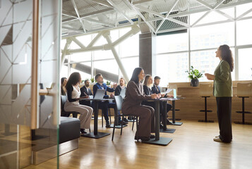 Speaker leads training seminar for office team. Engaged audience in a meeting listens to a coach giving a presentation in a bright modern workspace. Clear concept of team learning workshop.